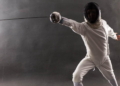 Boy wearing white fencing costume and black fencing mask standing with the sword practicing in fencing.