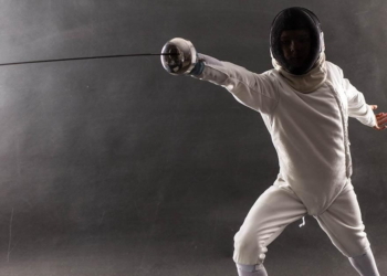 Boy wearing white fencing costume and black fencing mask standing with the sword practicing in fencing.