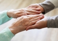 Mature female in elderly care facility gets help from hospital personnel nurse. Senior woman w/ aged wrinkled skin & care giver, hands close up. Grand mother everyday life. Background, copy space.