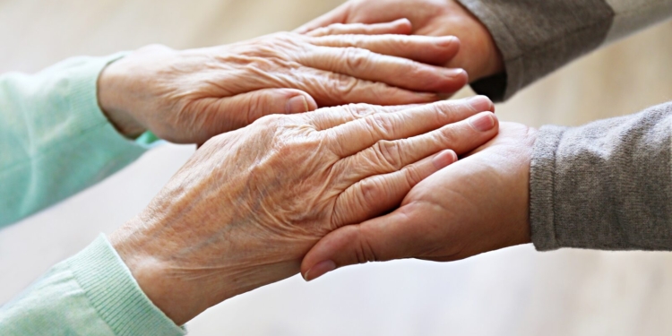 Mature female in elderly care facility gets help from hospital personnel nurse. Senior woman w/ aged wrinkled skin & care giver, hands close up. Grand mother everyday life. Background, copy space.